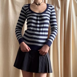 Puede incluir: Un su&eacute;ter de manga larga a rayas azul marino y gris con cuello redondo se combina con una minifalda plisada negra. El conjunto se completa con calcetines hasta la rodilla grises y tacones negros de punta. La ropa se muestra sobre un fondo neutro.