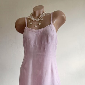 May include: A light pink slip dress featuring thin spaghetti straps, a delicate lace trim along the neckline, and a matching lace border detail at the hem. The garment is displayed on a mannequin, which is also wearing a white floral necklace.