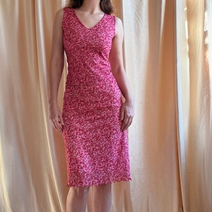 May include: A sleeveless, two-piece floral dress in shades of pink and red. The top has a V-neck, and the skirt is knee-length with a ruffled hem. The model is wearing green shoes. The background is a light beige curtain.