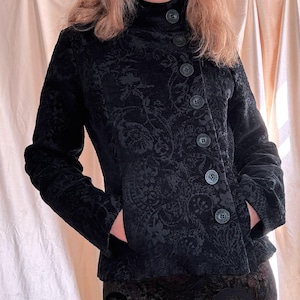 May include: A black jacket featuring a tonal floral jacquard pattern, a high collar, and a row of dark buttons running diagonally down the front. The jacket includes long sleeves and side pockets, paired with a matching black floral skirt.