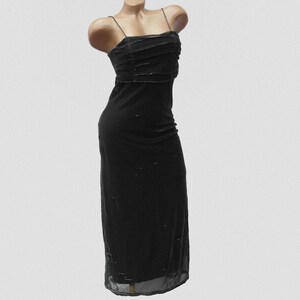 May include: A floor-length black dress featuring thin spaghetti straps and a straight neckline. The bodice is detailed with horizontal pleated fabric layers, while the skirt portion has a sheer overlay with subtle, scattered metallic accents.