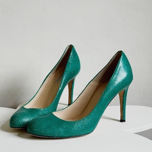 May include: A pair of teal green high-heeled shoes. The shoes have a textured, snakeskin-like pattern and a pointed toe. The heels are slender and the shoes are displayed on a white surface.