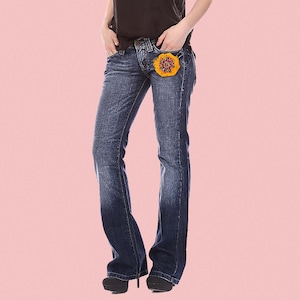 Może przedstawiać: Dżinsy w kolorze niebieskim z ozdobą w kształcie kwiatu na prawej kieszeni.