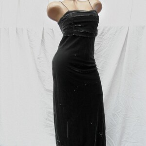 May include: A floor-length black dress featuring thin spaghetti straps and a layered bodice with horizontal detailing. The garment has a straight silhouette and includes scattered small embellishments throughout the fabric of the skirt.