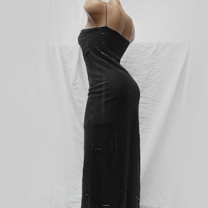 May include: A floor-length black dress featuring thin spaghetti straps and a fitted silhouette. The fabric has a subtle, semi-sheer texture with faint horizontal detailing throughout the garment, extending from the bodice down to the hemline.