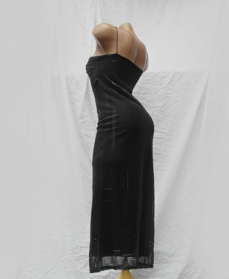 May include: A floor-length black dress featuring thin spaghetti straps and a fitted silhouette. The fabric has a subtle, semi-sheer texture with faint horizontal detailing throughout the garment, extending from the bodice down to the hemline.