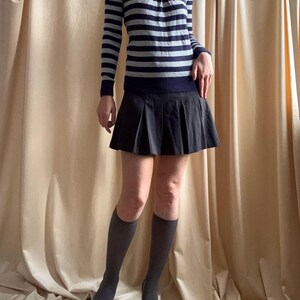 Puede incluir: Un su&eacute;ter de manga larga a rayas en azul marino y gris, combinado con una minifalda plisada negra. El conjunto se completa con calcetines hasta la rodilla grises y tacones negros de punta. Tambi&eacute;n se ve un collar con un colgante.