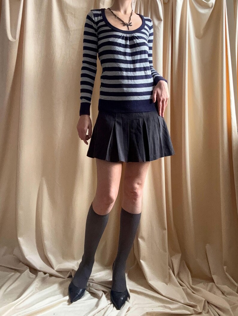 Puede incluir: Un su&eacute;ter de manga larga a rayas en azul marino y gris, combinado con una minifalda plisada negra. El conjunto se completa con calcetines hasta la rodilla grises y tacones negros de punta. Tambi&eacute;n se ve un collar con un colgante.