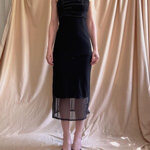 May include: A black midi-length dress featuring thin spaghetti straps and a bodice with horizontal velvet-like textured bands. The skirt section includes a sheer, semi-transparent mesh overlay at the hemline, creating a layered appearance.