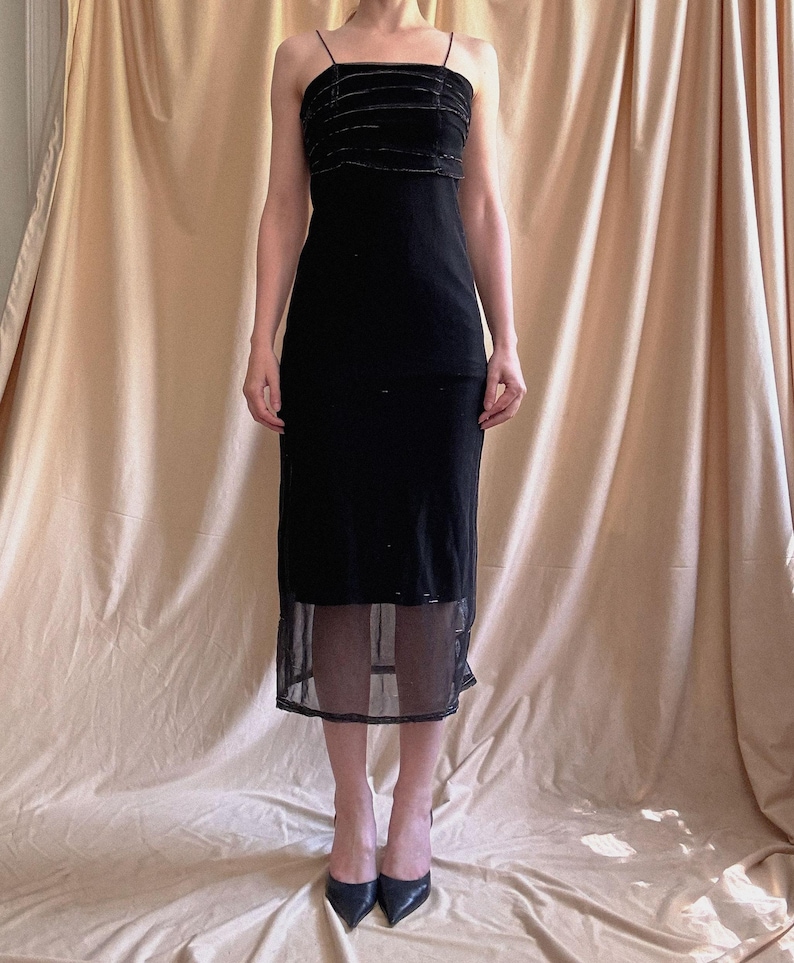 May include: A black midi-length dress featuring thin spaghetti straps and a bodice with horizontal velvet-like textured bands. The skirt section includes a sheer, semi-transparent mesh overlay at the hemline, creating a layered appearance.