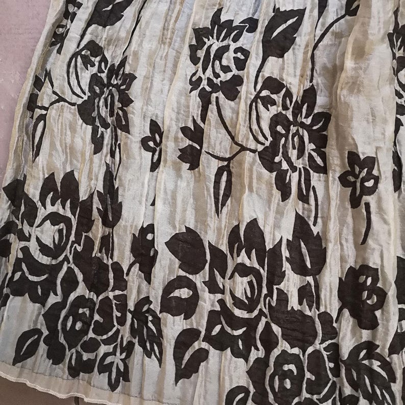 May include: A cream-colored skirt with a dark brown floral pattern. The skirt features a repeating design of large flowers and leaves, creating a bold contrast against the light background. The fabric appears to be lightweight and slightly textured.