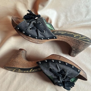 May include: A pair of wooden-soled sandals with denim uppers. The sandals feature a lace-up design, silver studs, and a small heel. The soles are brown with the word "Renley" printed on the side. A black tassel is on the top of the sandal.