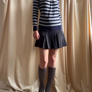 Puede incluir: Un su&eacute;ter de manga larga con cuello redondo y rayas horizontales azul marino y gris. El su&eacute;ter se combina con una falda plisada negra, calcetines hasta la rodilla grises y tacones negros puntiagudos. El conjunto se presenta sobre un fondo de tonos neutros.