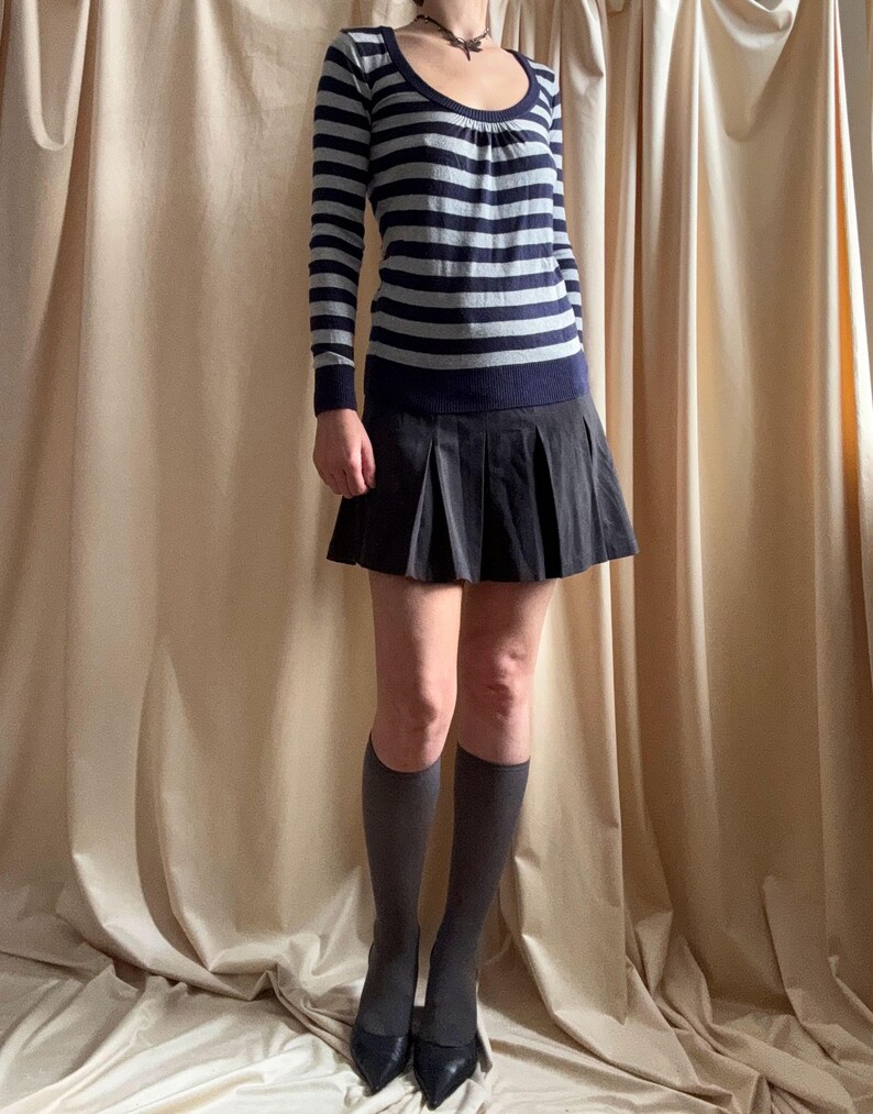 Puede incluir: Un su&eacute;ter de manga larga con cuello redondo y rayas horizontales azul marino y gris. El su&eacute;ter se combina con una falda plisada negra, calcetines hasta la rodilla grises y tacones negros puntiagudos. El conjunto se presenta sobre un fondo de tonos neutros.