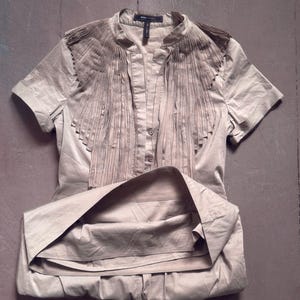 May include: A beige short-sleeved blouse with a mandarin collar and decorative fringe detail across the chest. The blouse is buttoned down the front and has a folded skirt. The fabric has a slight sheen.