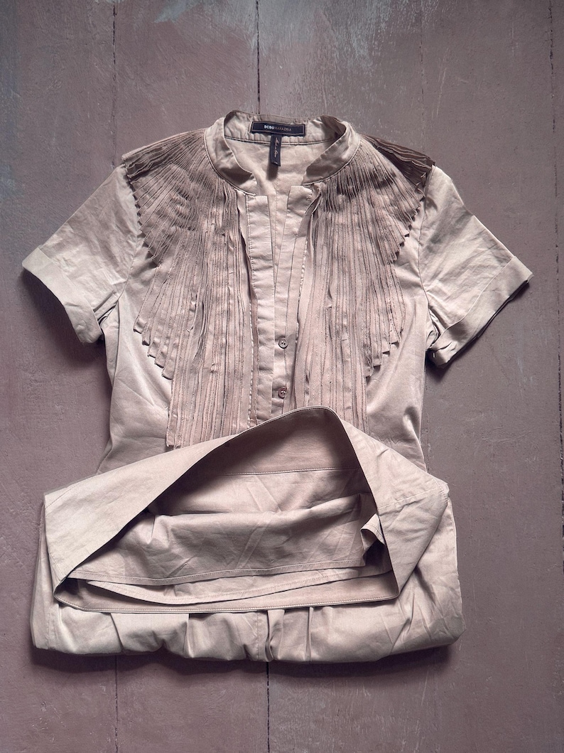 May include: A beige short-sleeved blouse with a mandarin collar and decorative fringe detail across the chest. The blouse is buttoned down the front and has a folded skirt. The fabric has a slight sheen.