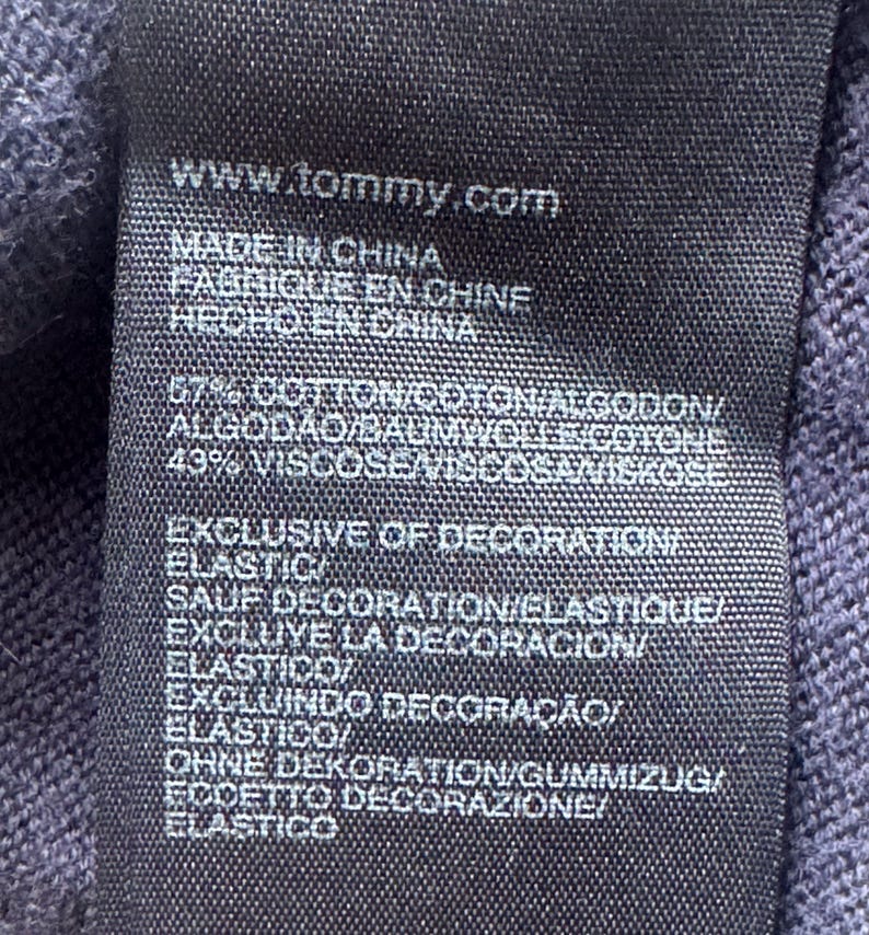 Puede incluir: Primer plano de una etiqueta de ropa azul marino oscuro con texto blanco. La etiqueta incluye la direcci&oacute;n del sitio web www.tommy.com y frases en varios idiomas, incluyendo "Made in China" y "Exclusive of Decoration".