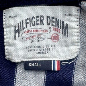 Puede incluir: Primer plano de una etiqueta de una prenda Hilfiger Denim. La etiqueta blanca tiene texto negro, incluyendo "HILFIGER DENIM 1985" y "NEW YORK CITY N.Y.C. UNITED STATES OF AMERICA." Una etiqueta m&aacute;s peque&ntilde;a dice "SMALL" con rayas rojas, blancas y azules.