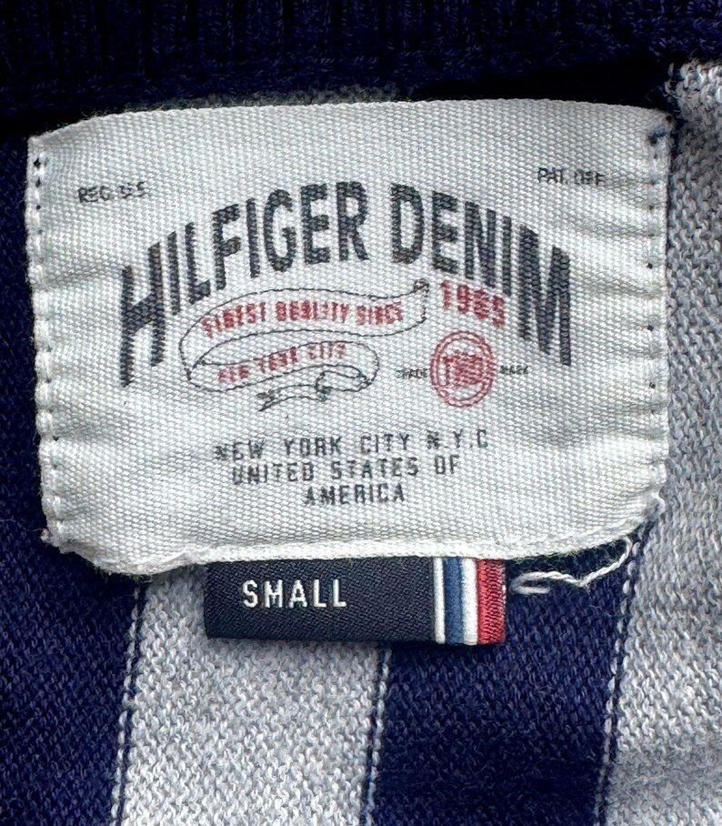Puede incluir: Primer plano de una etiqueta de una prenda Hilfiger Denim. La etiqueta blanca tiene texto negro, incluyendo "HILFIGER DENIM 1985" y "NEW YORK CITY N.Y.C. UNITED STATES OF AMERICA." Una etiqueta m&aacute;s peque&ntilde;a dice "SMALL" con rayas rojas, blancas y azules.