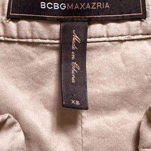 May include: Close-up of a tan garment with a black label that reads "BCBGMAXAZRIA" in white lettering. Below the label is a black tag that says "Made in China" and "XS".