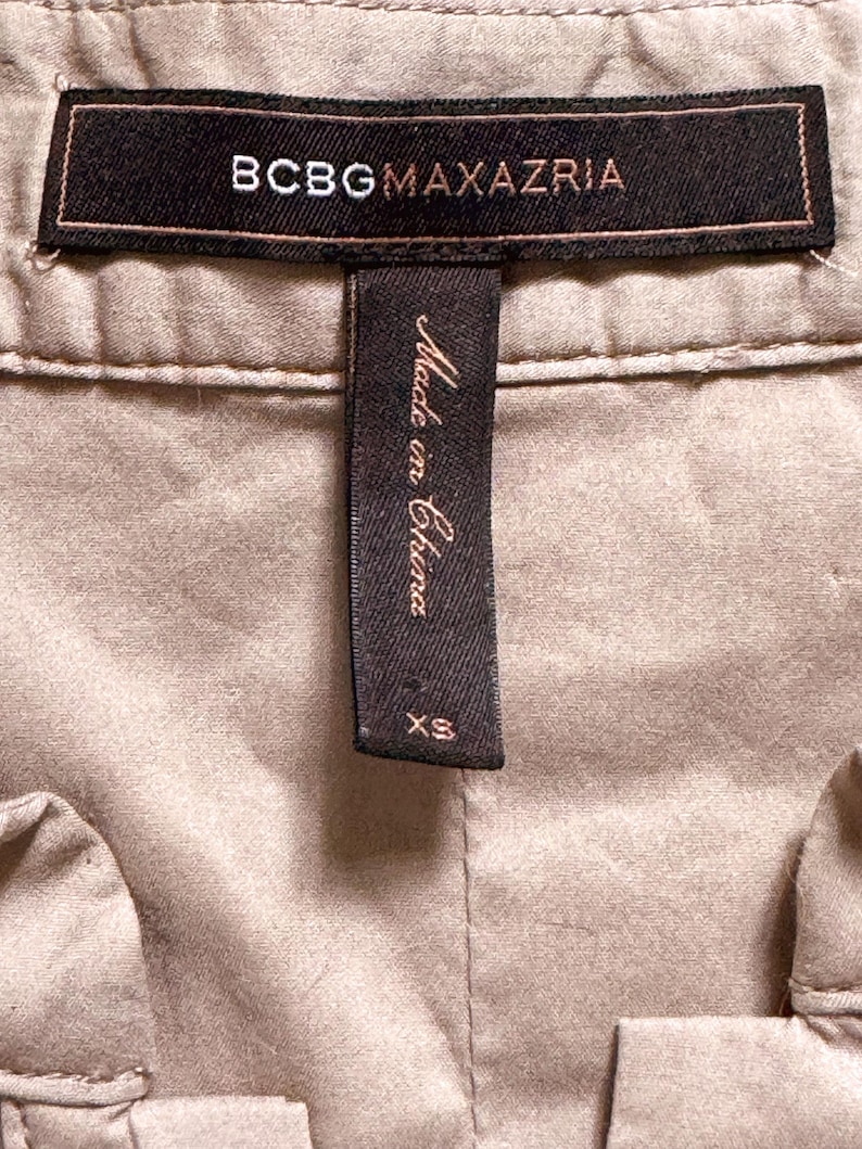 May include: Close-up of a tan garment with a black label that reads "BCBGMAXAZRIA" in white lettering. Below the label is a black tag that says "Made in China" and "XS".