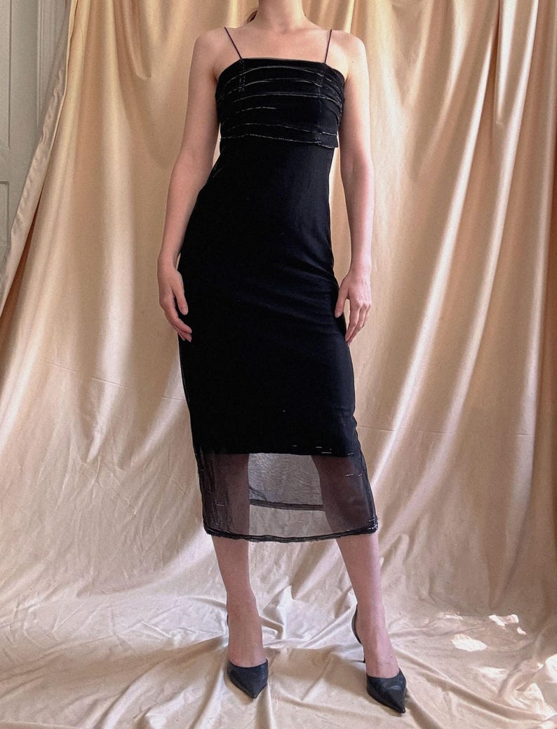 May include: A black midi-length dress featuring thin spaghetti straps and a square neckline. The bodice is accented with horizontal velvet-like bands, while the lower portion of the skirt transitions into a sheer, semi-transparent black mesh hem.