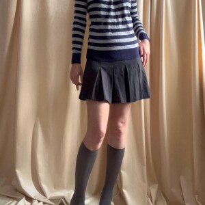 Puede incluir: Un jersey de manga larga a rayas azul marino y gris claro con cuello redondo se combina con una minifalda plisada negra. El conjunto se completa con calcetines hasta la rodilla grises y tacones negros de punta.