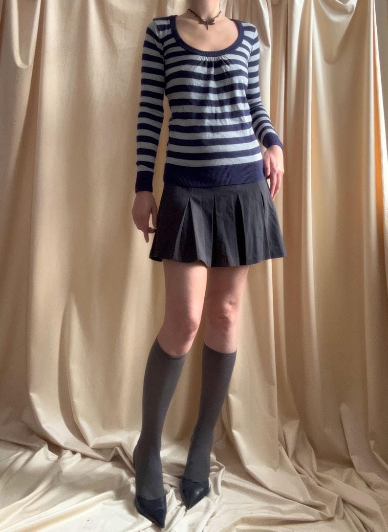 Puede incluir: Un jersey de manga larga a rayas azul marino y gris claro con cuello redondo se combina con una minifalda plisada negra. El conjunto se completa con calcetines hasta la rodilla grises y tacones negros de punta.