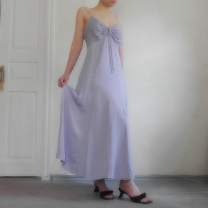 May include: A light purple maxi dress with spaghetti straps and a bow detail at the bust. The dress has a flowing skirt and is paired with black heeled sandals. The dress is made of a lightweight fabric.