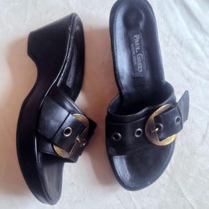 Può includere: Un paio di sandali con zeppa in pelle nera con grandi fibbie color oro. I sandali hanno una suola platform e il marchio "PAUL GREEN MUNCHEN" è visibile sulla soletta.