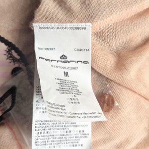 以下が含まれることがあります： 桃色の衣服のクローズアップ。タグにはブランド名「Fornarina」、サイズ「M」、および複数の言語でのケア手順が表示されています。小さくて丸い桃色のボタンが見えます。