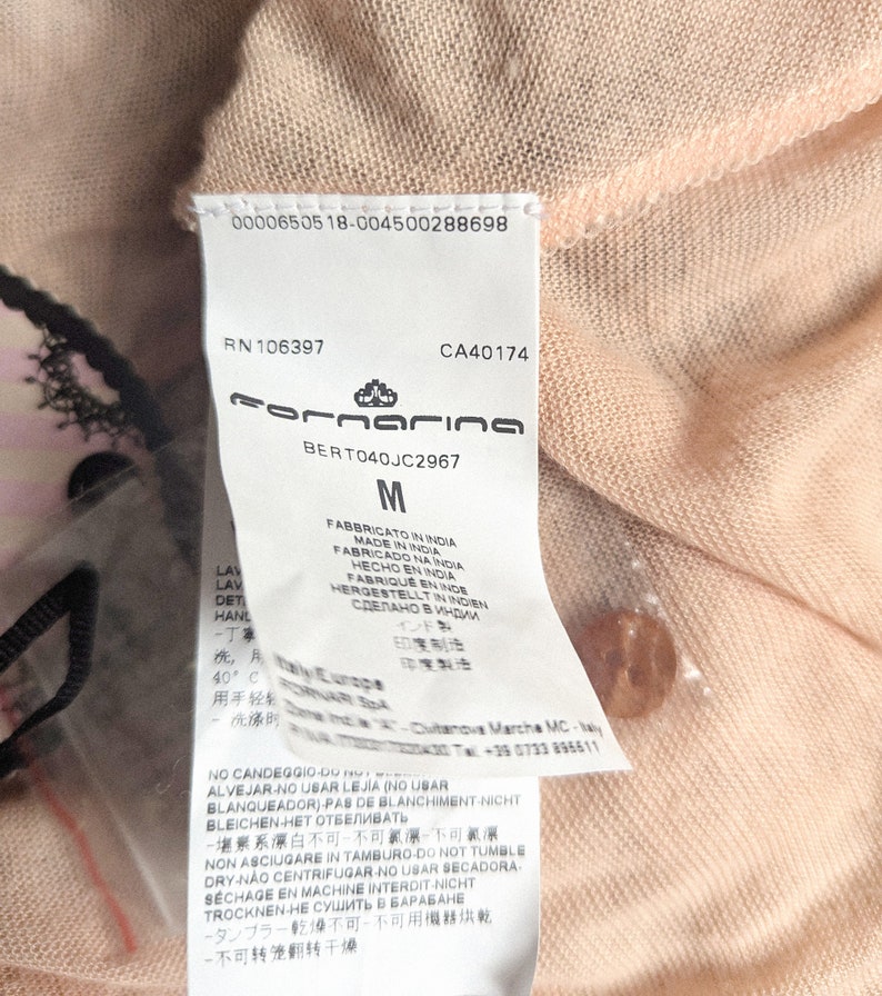 以下が含まれることがあります： 桃色の衣服のクローズアップ。タグにはブランド名「Fornarina」、サイズ「M」、および複数の言語でのケア手順が表示されています。小さくて丸い桃色のボタンが見えます。