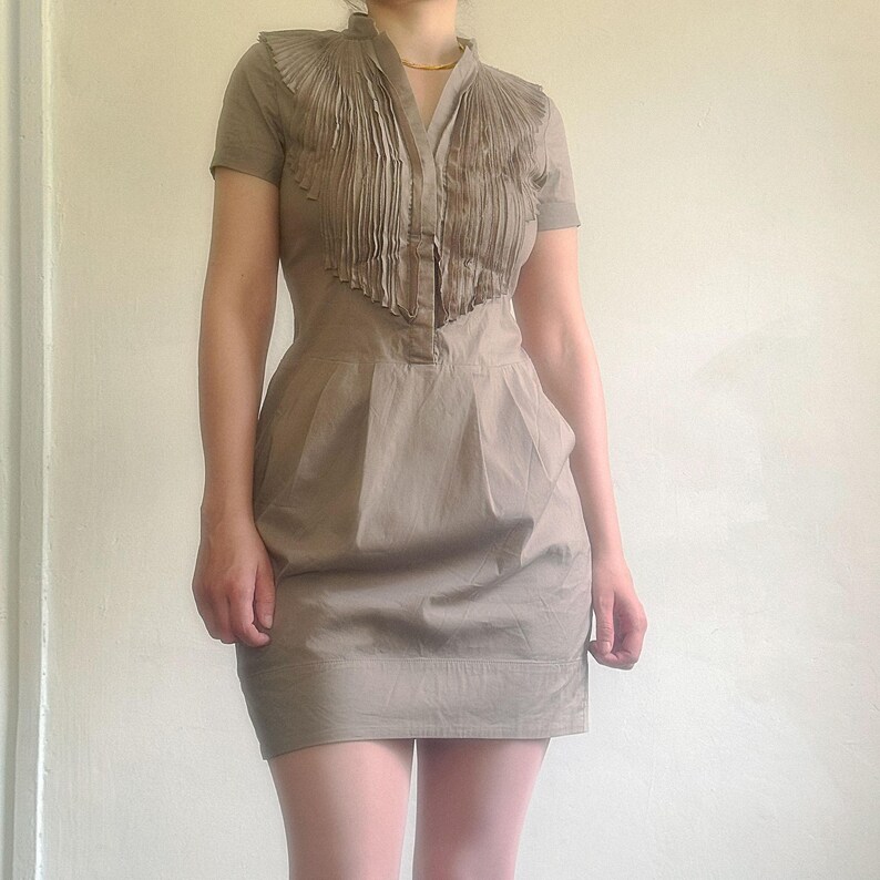 May include: A short-sleeved, taupe dress with a pleated bib detail and a V-neckline. The dress has a fitted bodice and a flared skirt, with pockets. The dress is made of a lightweight fabric and is suitable for warm weather.