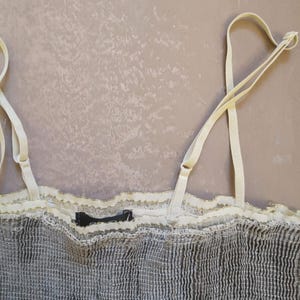 May include: A close-up of a gray camisole with adjustable cream-colored straps. The top features a textured fabric with a cream-colored lace trim along the neckline. A small black tag is visible.