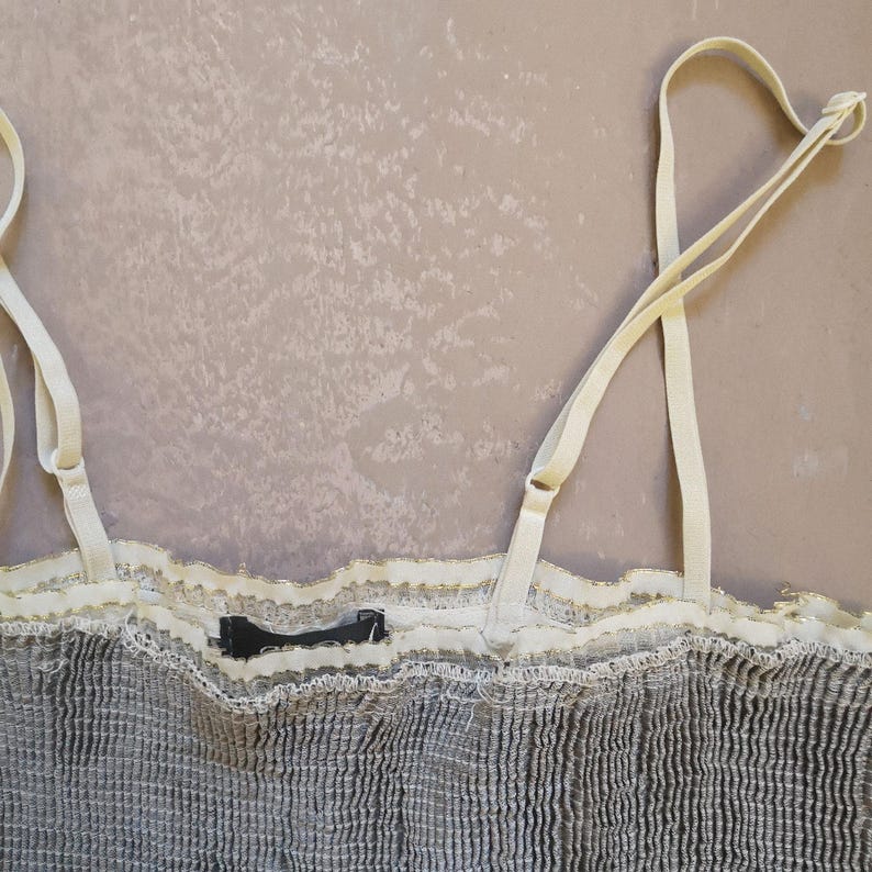May include: A close-up of a gray camisole with adjustable cream-colored straps. The top features a textured fabric with a cream-colored lace trim along the neckline. A small black tag is visible.