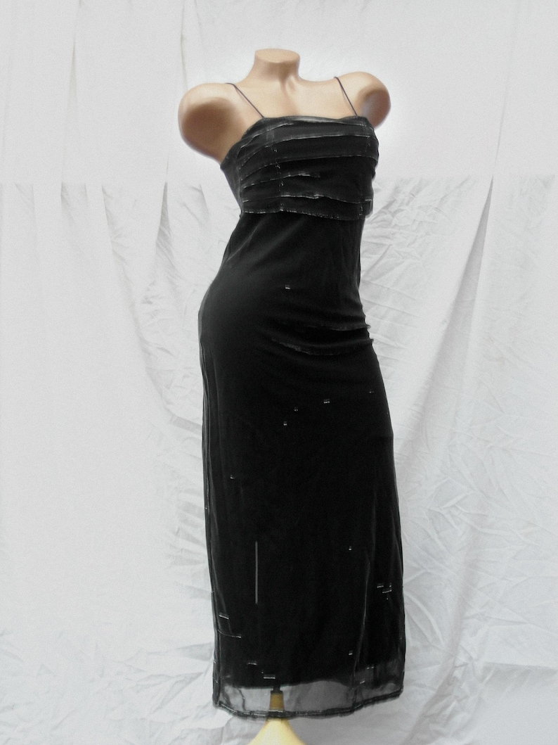 May include: A floor-length black dress featuring thin spaghetti straps and a layered bodice with horizontal detailing. The garment has a straight silhouette and includes scattered small embellishments throughout the fabric of the skirt.