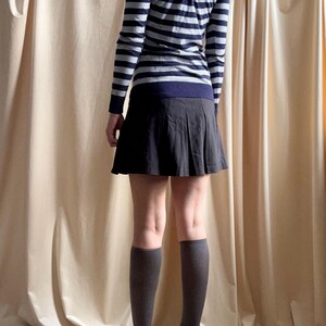 Puede incluir: Un su&eacute;ter de manga larga a rayas azul marino y gris con un detalle de ojo de cerradura en la espalda. El conjunto incluye una falda plisada gris oscuro, calcetines hasta la rodilla grises y zapatos de tac&oacute;n alto negros. La ropa se muestra sobre un fondo de tonos neutros.