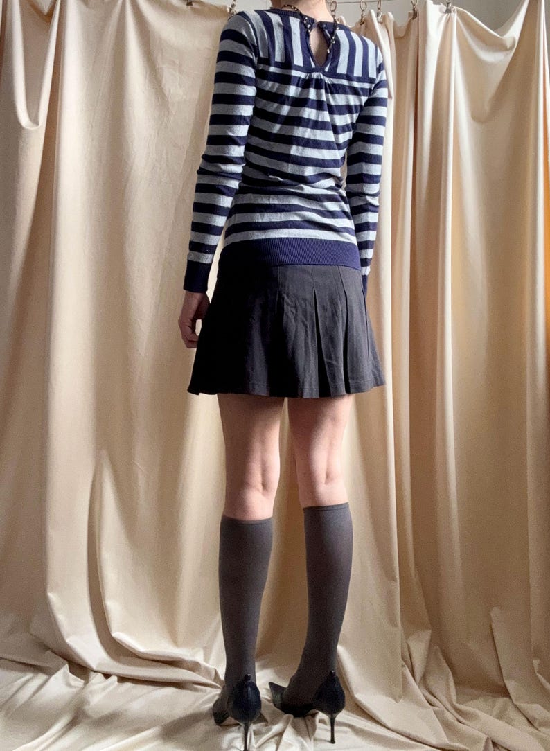 Puede incluir: Un su&eacute;ter de manga larga a rayas azul marino y gris con un detalle de ojo de cerradura en la espalda. El conjunto incluye una falda plisada gris oscuro, calcetines hasta la rodilla grises y zapatos de tac&oacute;n alto negros. La ropa se muestra sobre un fondo de tonos neutros.