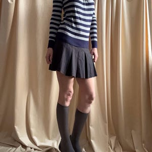 Puede incluir: Un su&eacute;ter de manga larga con cuello redondo y rayas horizontales azul marino y gris. Combinado con una minifalda plisada gris oscuro, calcetines hasta la rodilla y tacones negros. El conjunto se muestra sobre un fondo de tonos neutros.