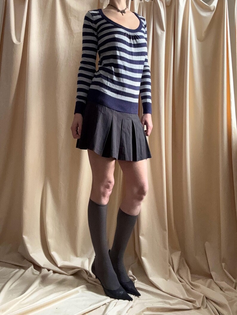 Puede incluir: Un su&eacute;ter de manga larga con cuello redondo y rayas horizontales azul marino y gris. Combinado con una minifalda plisada gris oscuro, calcetines hasta la rodilla y tacones negros. El conjunto se muestra sobre un fondo de tonos neutros.