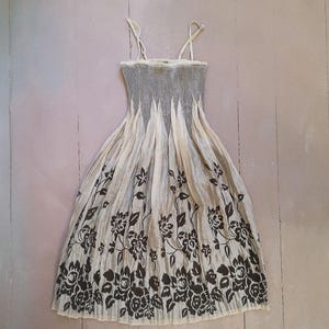 May include: A cream-colored sundress with a gray bodice and floral print. The dress features thin straps and a pleated skirt with a dark brown floral pattern along the bottom. The dress is made of a lightweight fabric.
