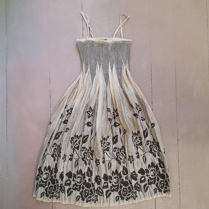 May include: A cream-colored sundress with a gray bodice and floral print. The dress features thin straps and a pleated skirt with a dark brown floral pattern along the bottom. The dress is made of a lightweight fabric.