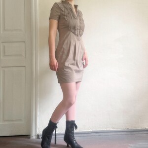 May include: A tan-colored dress with short sleeves and a collared neckline, featuring fringe detailing across the chest. The dress has a fitted silhouette and is paired with black ankle boots. The boots have a lace-up design and small heels.
