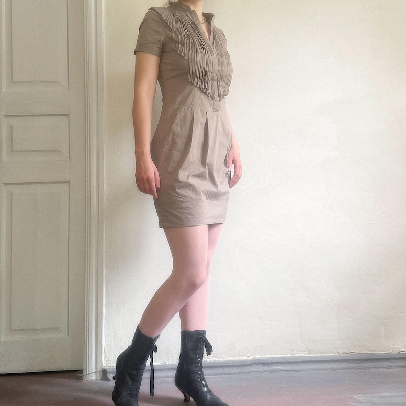 May include: A tan-colored dress with short sleeves and a collared neckline, featuring fringe detailing across the chest. The dress has a fitted silhouette and is paired with black ankle boots. The boots have a lace-up design and small heels.