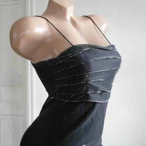 May include: A black dress featuring thin spaghetti straps and a bodice with horizontal pleated detailing. The fabric has a subtle metallic sheen, and the garment is displayed on a mannequin to show the fitted silhouette and the textured design.