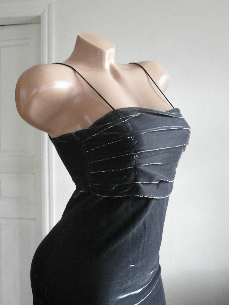 May include: A black dress featuring thin spaghetti straps and a bodice with horizontal pleated detailing. The fabric has a subtle metallic sheen, and the garment is displayed on a mannequin to show the fitted silhouette and the textured design.