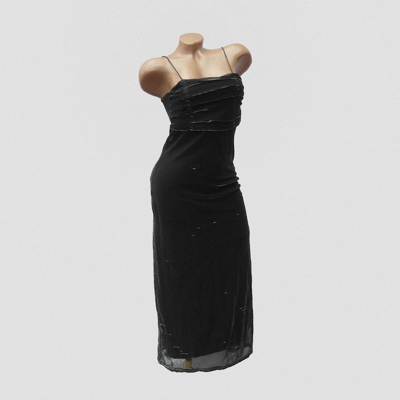 May include: A floor-length black dress featuring thin spaghetti straps and a straight neckline. The bodice is detailed with horizontal pleated fabric layers, while the skirt portion has a sheer overlay with subtle, scattered metallic accents.