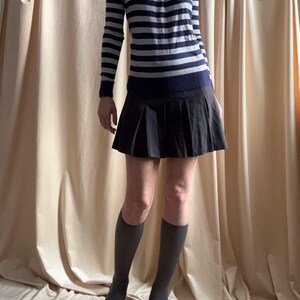 Puede incluir: Un jersey de manga larga a rayas azul marino y gris con cuello redondo se combina con una falda plisada negra. El conjunto se completa con calcetines altos grises hasta la rodilla y tacones negros de punta. La ropa se muestra sobre un fondo neutro.