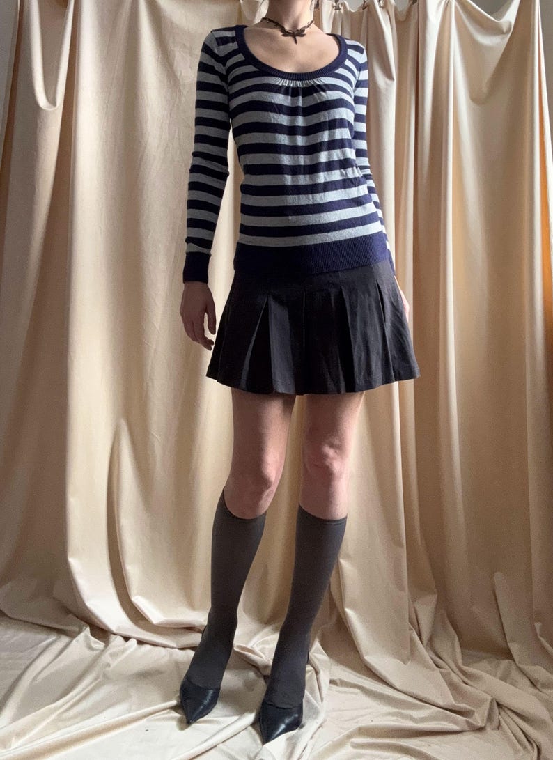 Puede incluir: Un jersey de manga larga a rayas azul marino y gris con cuello redondo se combina con una falda plisada negra. El conjunto se completa con calcetines altos grises hasta la rodilla y tacones negros de punta. La ropa se muestra sobre un fondo neutro.