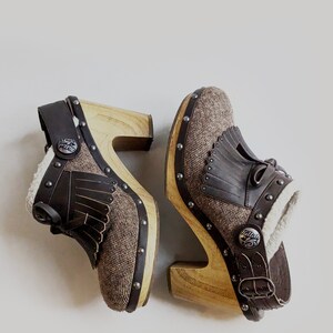 May include: A pair of brown and beige suede clogs with wooden heels. The clogs have a fringe detail and a buckle closure. The clogs are decorated with metal studs and a metal tag that says "Buffalo".